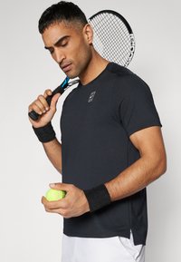 Svart treningstrøye med korte ermer og rund hals, kombinert med et svart håndleddband; holder en lys grønn tennisball.