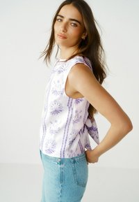Ung kvinde med langt brunt hår if�ørt en ærmeløs hvid top med lilla blomsterprint og lyseblå jeans, der poserer sidelæns mod en ensfarvet baggrund.