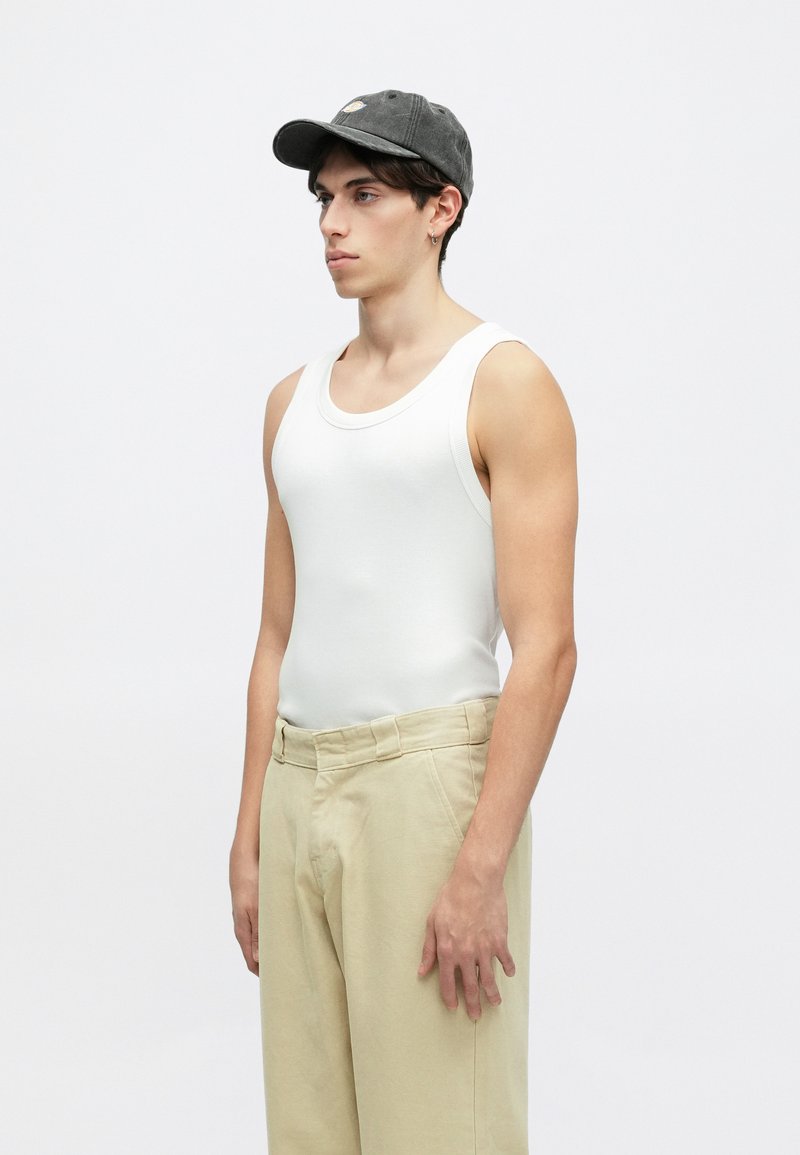 Canotta bianca a costine, pantaloni crema e berretto grigio scuro con logo. Il modello è in posizione laterale su uno sfondo bianco.