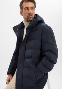 Marineblauwe pufferjas met capuchon, met een glad, doorgestikt textuur. Voorzien van een ronde logo op de mouw en een knoopsluiting aan de voorkant.