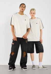 Deux individus portent des t-shirts crèmes surdimensionnés avec des accents noirs rayés. La personne à gauche a des jeans noirs déchirés ; l'autre porte un short noir.