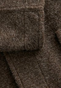 Tissu en laine marron présentant une surface texturée et duveteuse avec des fibres visibles, des coutures piquées et un bord replié, mettant en valeur sa douceur et sa chaleur.