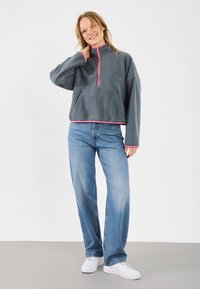 Grauer Fleecepullover mit einem halben Reißverschluss am Kragen, versehen mit rosa Akzenten und einer vorderen Kängurutasche. Kombiniert mit hellblauen Jeans mit weitem Bein.