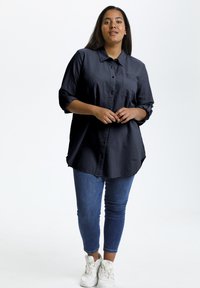 Chemise bleu foncé à manches retroussées, en tissu texturé, avec une poche poitrine, associée à un jean skinny bleu et des baskets blanches.