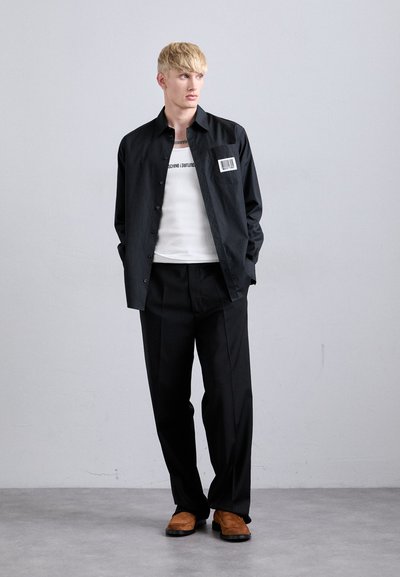 Camisa preta com botões e um patch de código de barras, top branco de alças, calças pretas ajustadas e sapatos castanhos, tudo sobre um fundo neutro.