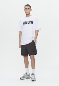 T-shirt blanche avec impression noire "HUMAN" et shorts noirs. Le modèle porte des baskets grises et des chaussettes blanches. Tenue simple et décontractée avec des couleurs neutres.