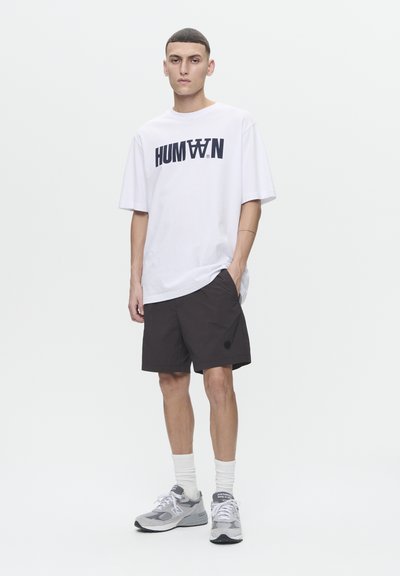 T-shirt blanche avec impression noire "HUMAN" et shorts noirs. Le modèle porte des baskets grises et des chaussettes blanches. Tenue simple et décontractée avec des couleurs neutres.