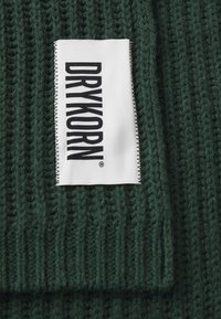 Knitted green fabric with a ribbed texture. A white label features the brand name "DRYKORN" in black bold letters, stitched on the edge.
