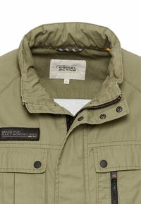 Olive green jacket with a zip front, two chest pockets, and a text patch. Features a stand collar with a checkered lining and snap buttons.
