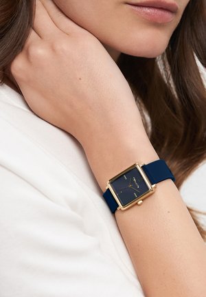 Goldfarbene rechteckige Uhr mit einem nav blauen Silikonarmband. Das Zifferblatt ist dunkelblau und verfügt über minimalistische Stundenmarkierungen.