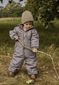 Szary wodoodporny kombinezon śnieżny z zamkiem błyskawicznym z przodu, elastycznymi mankietami i kapturem. Wnętrze wyłożone brązową tkaniną. Brązowe buty; dziecko trzyma kij na trawie.