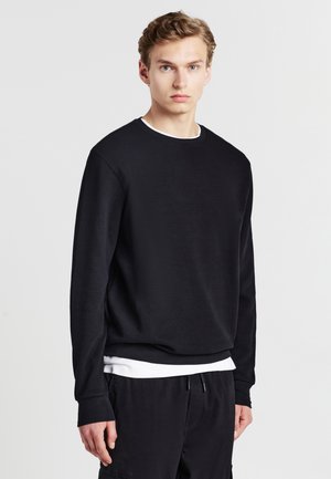 Sweatshirt noir à manches longues avec une finition texturée, encolure ronde et poignets côtelés, superposé sur une chemise blanche, assorti à un pantalon noir.