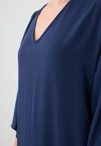Top blu navy con scollatura a V e vestibilità ampia, realizzato in tessuto testurizzato, dotato di maniche a 3/4 per una maggiore libertà di movimento.