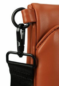 Eckausschnitt einer braunen strukturierten Ledertasche mit einem schwarzen Metallclip und Riemen. Ein runder Ring verbindet den Clip mit der Tasche und hebt das funktionale Design hervor.
