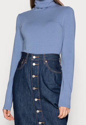 Mujer con un suéter de cuello alto de manga larga azul claro metido en una falda de mezclilla oscura de talle alto con botones al frente, con la mano relajada al costado.