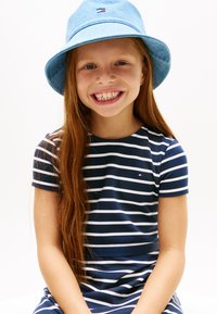 Χαμογελαστή κοπέλα με μακριά κόκκινα μαλλιά, φορούν μπλε και άσπρο ριγέ φόρεμα και ανοιχτό μπλε καπέλο τύπου κουβά, πάνω σε λευκό φόντο.