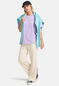 Lavendel t-shirt, lyseblå sweater draperet over skuldrene, cremfarvede bukser med brede ben og sorte sneakers med hvide såler. Simpel, afslappet design.