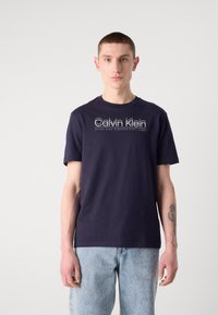Marineblauwe katoenen t-shirt met een witte Calvin Klein logo-print op de borst, korte mouwen en een ronde hals. Model draagt spijkerbroek.