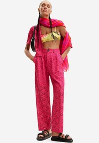 Fuchsia kanten broek met een losse pasvorm en een gedetailleerd bloemmotief, gecombineerd met een gele crop top met patroon en een roze sjaal. Zwarte sandalen.