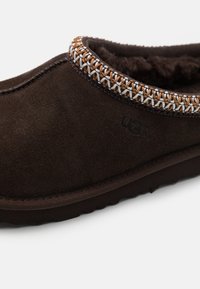 Pantoufle en daim marron foncé avec une doublure en laine, ornée d'accents brodés marron et blanc autour du col, et d'une semelle en caoutchouc texturé.