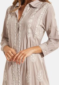 Chemise boutonnée gris clair à manches longues, avec broderie florale blanche, texture plissée et col classique. Tissu doux.