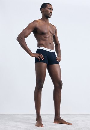 Černé pánské boxerky s bílým lemováním, přiléhavý střih a bavlněný materiál. Postojící model na neutrálním pozadí.