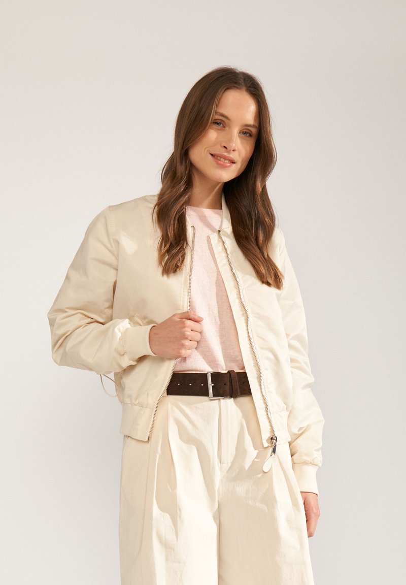 Femme aux longs cheveux bruns portant une veste bomber beige, une chemise rose clair, un pantalon large crème et une ceinture marron foncé, souriant légèrement.