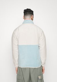 Giacca in fleece con una parte superiore color crema e una parte inferiore azzurro chiaro. Dettagli con colletto alto e tessuto testurizzato. Cuciture rinforzate.