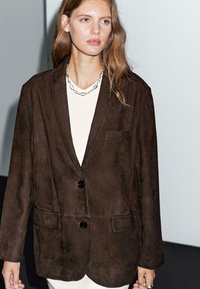 Blazer en cuir suédé marron avec une coupe décontractée, deux poches avant et deux boutons noirs. Porté par-dessus un haut de couleur claire. Design simple et texturé.