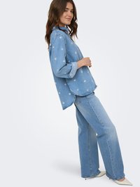 Mujer con una camisa vaquera azul con patrones de flores blancas y vaqueros anchos de pierna ancha azul claro, de pie de lado con las manos cerca de la cintura.