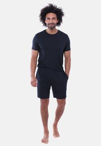 T-shirt nera a maniche corte con tasca e pantaloncini neri abbinati. Il tessuto è liscio, con una vestibilità comoda. Il modello è in piedi a piedi nudi su uno sfondo semplice.