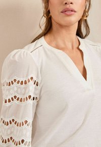 White blouse with a V-neck, featuring puff sleeves and intricate cut-out lace detailing. Soft texture and lightweight material.
