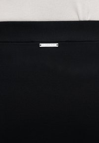 Κοντινή εικόνα μαύρης ζώνης υφάσματος με μια μικρή ορθογώνια ασημένια ετικέτα λογότυπου Michael Kors στο κέντρο κοντά στο πάνω άκρο.