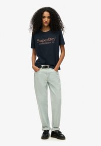 Marineblå bomulds t-shirt med "SuperDry Venice Beach, CA" logo, parret med lyseblå baggy jeans og sorte sko, samt et læderb�ælte.