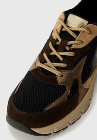 Sneaker featuring a mesh black upper, brown suede and leather accents, beige sole, and beige laces detailed with a metallic finish.