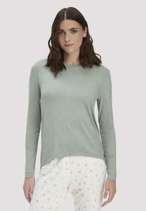 Junge Frau mit schulterlangem braunem Haar, die ein hellgrünes Langarmshirt und weiße Hosen mit kleinem orangefarbenem Blumenmuster trägt.