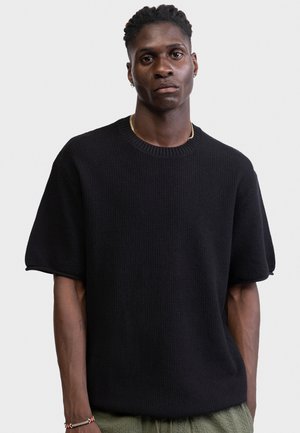 Νεαρός με κοντά σγουρά μαλλιά, φοράει μαύρο πλεκτό πουλόβερ με κοντά μανίκια και ελιάς πράσινο παντελόνι, μπροστά σε απλό φόντο.