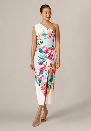 Vrouw staand, draagt een witte jurk met één schouder, met een levendige meerkleurige bloemenprint en een voorsplit, gecombineerd met metallic sandaalhakken met bandjes.