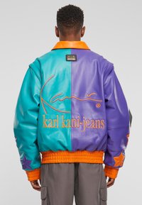 Kožená bombardérka s rozděleným designem v teal a fialové barvě, oranžovým límcem, žebrovanými manžetami a hvězdnými akcenty na rukávech s vyšívaným brandingem.