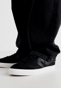 Schwarze Sneakers verfügen über ein strukturiertes Wildleder- und glattes Lederoberteil, eine weiße Gummisohle und eine Schnürdesign, kombiniert mit dunkelblauen Jeans.