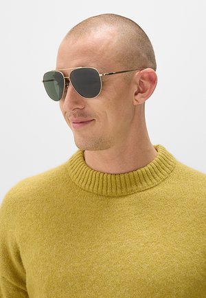 Uomo con testa rasata che indossa occhiali da sole aviator con montatura dorata e un maglione giallo senape, che guarda di lato con un leggero sorriso.