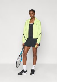 Haut à zip vert néon, débardeur noir, jupe noire, tenant une raquette de tennis et une balle. Chaussures de sport noires, chaussettes blanches, détails du logo mis en valeur.