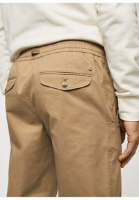 Pantalon kaki avec une taille élastique froncée, poches avant et arrière, accentué par de petits boutons en métal et des détails cousus.