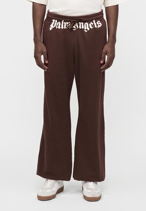 Personne portant un pantalon marron à jambes larges avec le texte "Palm Angels" sur la ceinture et des baskets blanches avec semelles en gomme.
