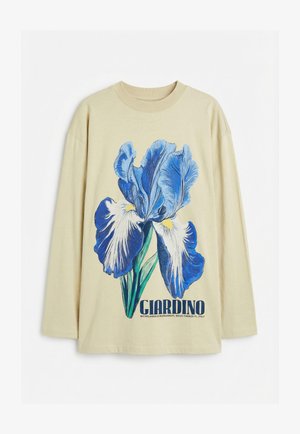 Beige lange mouwen shirt met grote blauwe en witte irisbloem grafiek en "GIARDINO" tekst gedrukt onder de bloem.