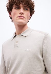 Νεαρός άνδρας με σγουρά μαλλιά, που φοράει ένα ανοιχτό μπεζ μακρυμάνικο πόλο με το λογότυπο "BOSS" στο στήθος, κοιτάζοντας ελαφρώς προς το πλάι.