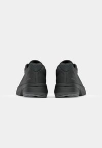 Des chaussures de sport noires avec une tige en mesh, un talon texturé et une semelle en caoutchouc épaisse. Présentent un branding minimal et un design épuré.