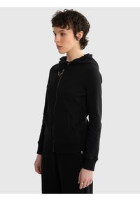 Persoană tânără cu păr scurt și creț, privind spre stânga, purtând un hanorac negru cu fermoar și buzunare frontale, și pantaloni negri, pe un fundal gri.