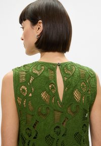 Πράσινο τοπ με δαντέλα που περιλαμβάνει περίπλοκα σχέδια με κοπές και κλείσιμο με κουμπί στην πλάτη. Ο σχεδιασμός περιέχει λουλούδια και κυματοειδή μοτίβα.