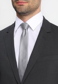 Grau Anzugjacke mit Revers, kombiniert mit einem weißen Hemd und einer strukturierten silbernen Krawatte, die ein dezentes Taschen-Detail aufweist.